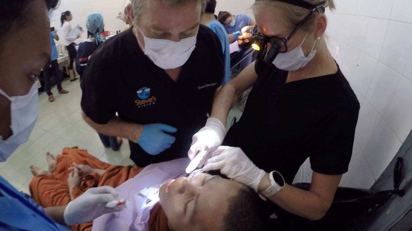 Steve Bender watches closely as a dentist with a headlamp treats a monk in orange robes