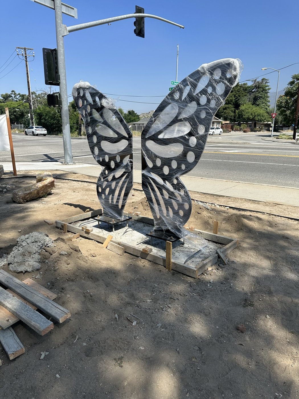 A large steel butterfly sculpture, still wrapped in plastic, being installed on a new concrete base at a street corner