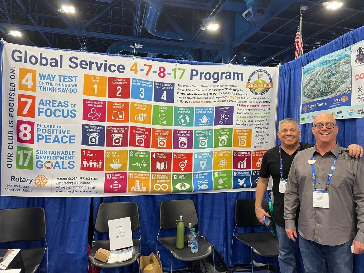 Steve and Don Bender stand beside the Rotary Club of Newport Beach Global Service 4-7-8-17 Program banner, with a Rotary Reefs poster behind them